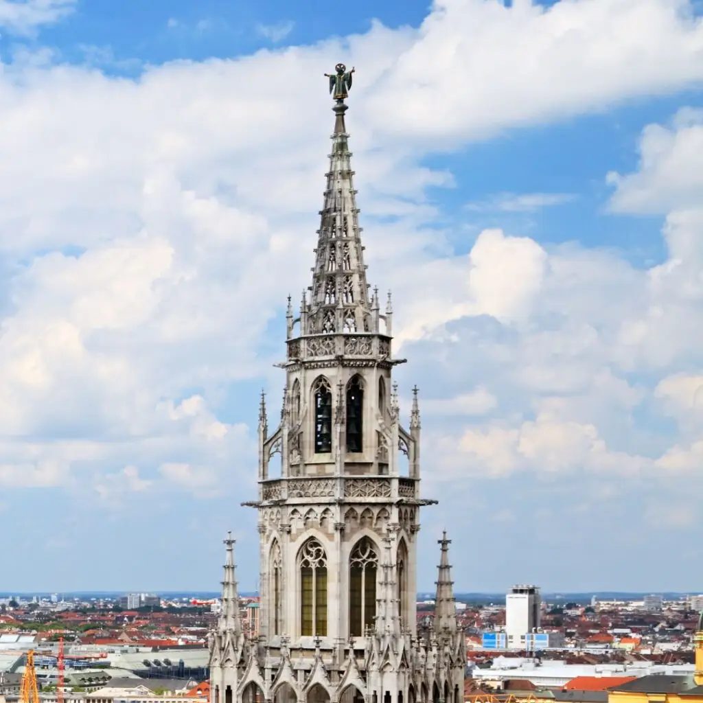 Spitze des Münchner Neuen Rathauses mit der Statue des „Patrona Bavariae“ vor leicht bewölktem Himmel. Die neugotische Architektur thront über der Altstadt – Symbol für Führung, Weitsicht und demokratischen Schulterschluss.