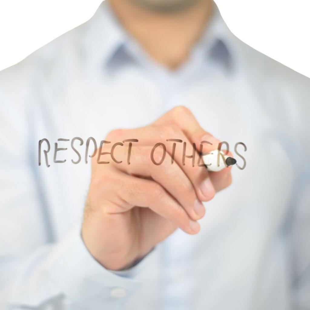 Ein Mann in einem hellblauen Hemd schreibt mit einem schwarzen Stift die Worte Respect Others auf eine durchsichtige Glasfläche. Der Fokus liegt auf der Hand und dem Schriftzug, der Respekt und Wertschätzung symbolisiert.