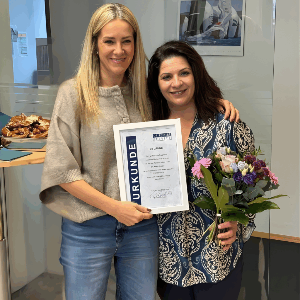 Constance Rettler, Geschäftsführerin Dr. Rettler Service Gebäudereinigung München steht gemeinsam mit der Jubiliarin im DRS-Büro und halten eine Jubiläumsurkunde und einen Blumenstrauß. Beide lächeln, im Hintergrund sind ein Kalender, ein Tisch mit Snacks und Glaselemente des Raumes zu sehen