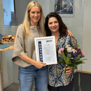 Constance Rettler, Geschäftsführerin Dr. Rettler Service Gebäudereinigung München steht gemeinsam mit der Jubiliarin im DRS-Büro und halten eine Jubiläumsurkunde und einen Blumenstrauß. Beide lächeln, im Hintergrund sind ein Kalender, ein Tisch mit Snacks und Glaselemente des Raumes zu sehen