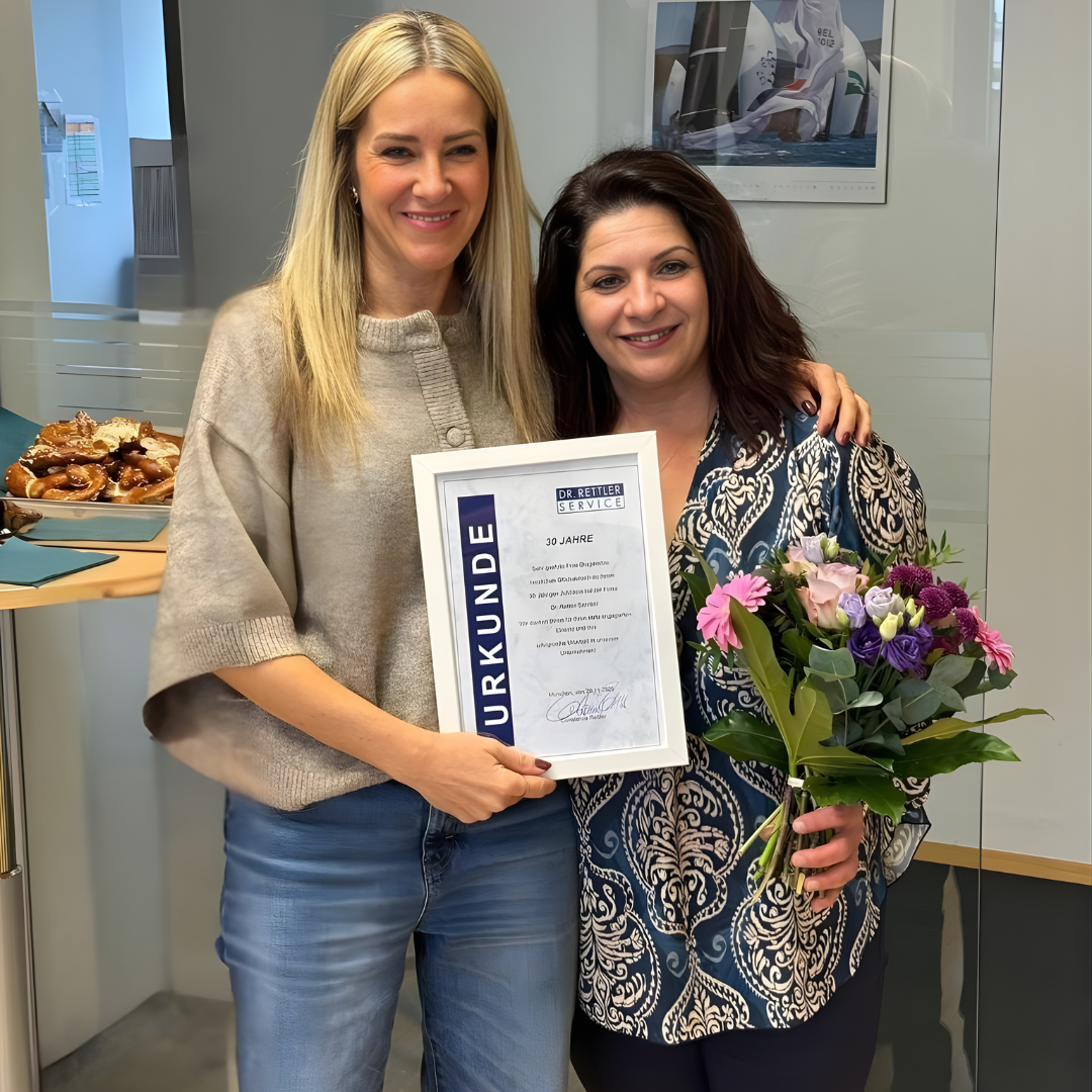 Constance Rettler, Geschäftsführerin Dr. Rettler Service Gebäudereinigung München steht gemeinsam mit der Jubiliarin im DRS-Büro und halten eine Jubiläumsurkunde und einen Blumenstrauß. Beide lächeln, im Hintergrund sind ein Kalender, ein Tisch mit Snacks und Glaselemente des Raumes zu sehen